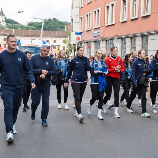 Eine Gruppe von Athleten in Team-Uniformen läuft bei einem Umzug die Straße hinunter. Sie wird von einem Mann vorne angeführt. Hinter ihnen sind Gebäude sichtbar.