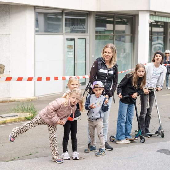 Eine Gruppe von Kindern und Erwachsenen, einige mit Helmen, stehen auf einer Stadtstraße, lächeln und posieren für ein Foto mit einem Gebäude im Hintergrund.