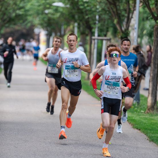 Mehrere Läufer nehmen an einem Marathon teil und tragen weiße und rote Uniformen. Sie laufen auf einem Betonweg, der von grünen Bäumen umgeben ist.
