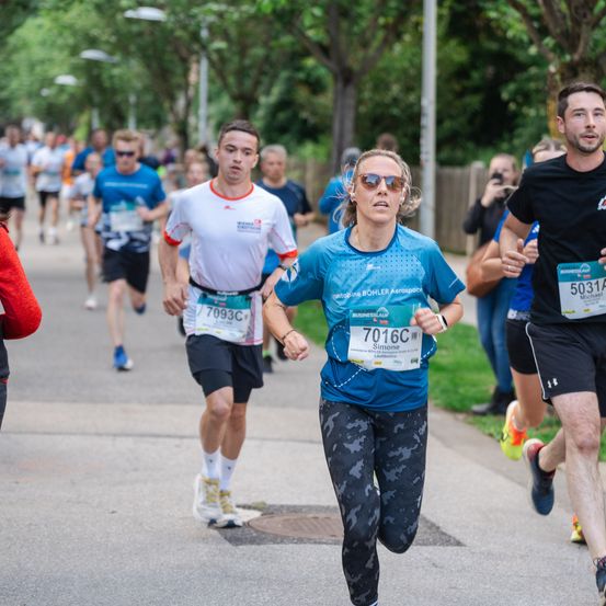 Eine Gruppe von Läufern nimmt an einem Marathon teil. Die Frau vorne trägt ein blaues Shirt und eine Sonnenbrille. Hinter ihr sind zwei Männer, einer trägt ein weißes Shirt. Der Weg hat Bäume auf beiden Seiten.