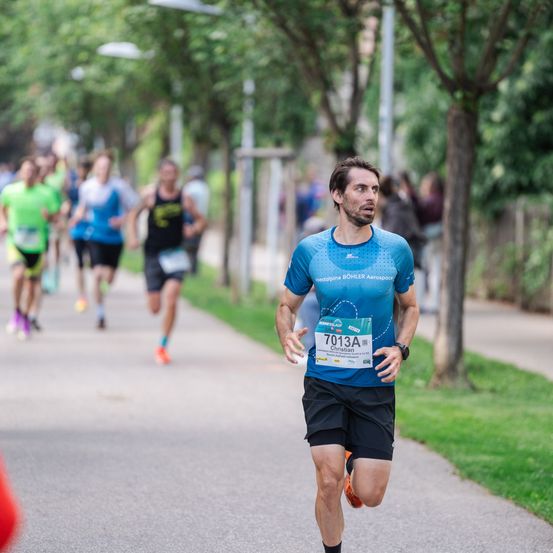 Ein Läufer in einem blauen Shirt und schwarzen Shorts läuft einen Marathon in der Stadt. Er trägt ein Startnummernband mit der Nummer 7013A. Hinter ihm folgen andere Läufer. Bäume und Gras säumen den Weg.