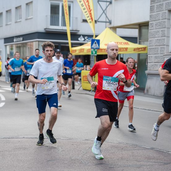 Mehrere Läufer nehmen an einem Marathon teil, jeder mit einer Startnummer. Der Mann in Rot, Nummer 7064C, führt das Feld an. Im Hintergrund sind ein gelbes Zelt und Gebäude zu sehen.