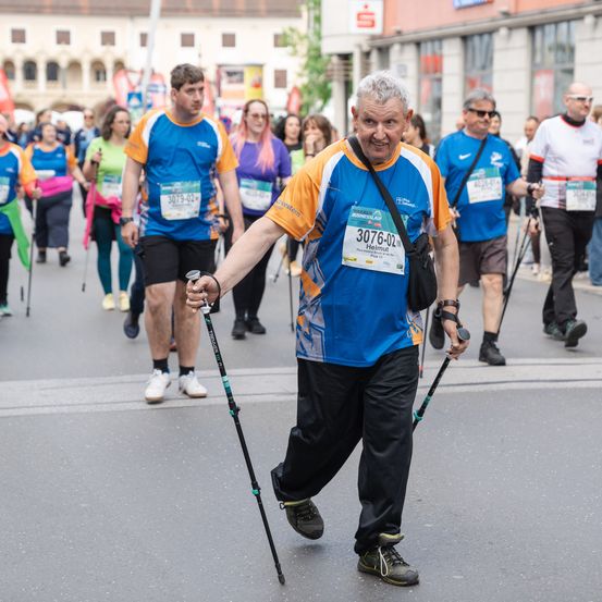 Ein älterer Mann in einem blauen T-Shirt mit der Nummer 3076 läuft in einem Marathon mit Trekkingstöcken. Hinter ihm laufen mehrere Teilnehmer ebenfalls mit Trekkingstöcken. Gebäude und Fahnen sind im Hintergrund.