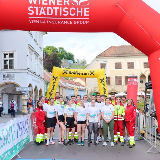 Eine Gruppe von Menschen in weißen Shirts mit Nummern auf ihnen posiert für ein Foto vor einem roten Bogen. Sie tragen grüne Hosen. Im Hintergrund ist ein Gebäude mit einem Schild Kleine Zeitung zu sehen.