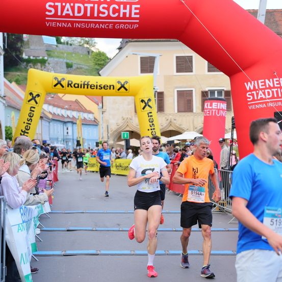 Marathonläufer überqueren die Ziellinie. Die Frau trägt Weiß, der Mann Orange. Zuschauer jubeln an der Seite. Werbung für Städtische und Raiffeisen ist sichtbar.