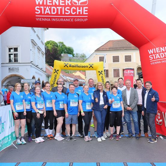 Eine Gruppe von Menschen in blauen T-Shirts steht vor einem roten Tor mit Text und einem gelben Tor. Sie alle lächeln und posieren für ein Foto. Das Tor sagt 'Wiener Städtische'.