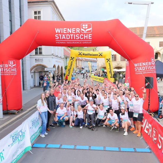 Eine Gruppe von Läufern posiert für ein Foto vor einem roten Bogen mit dem Logo Wiener Stadtische Vienna Insurance Group. Sie tragen alle weiße Shirts und einige tragen Turnschuhe.