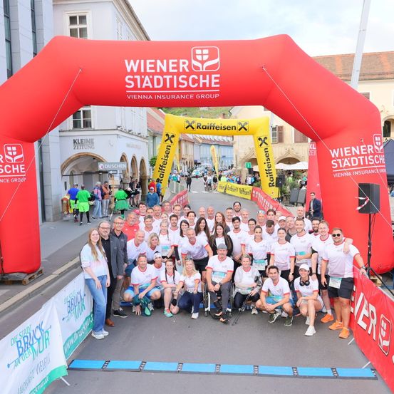 Eine Gruppe von Menschen posiert vor einem roten Bogen, gesponsert von Wiener Städtische. Im Hintergrund sind Gebäude und eine belebte Straße zu sehen.