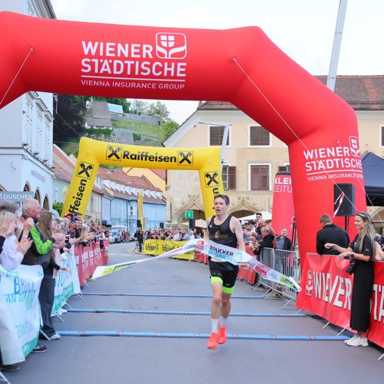 Ein Marathonläufer beendet das Rennen, durchquert einen rot-gelben Bogen und wird von Zuschauern angefeuert. Sponsorenbanner sind sichtbar.