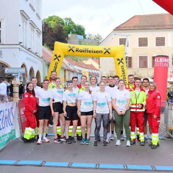Eine Gruppe von Menschen in übereinstimmenden weißen Shirts und Shorts steht unter einem gelben Banner bei einem Marathon. Eine Frau in Rot steht vorne.