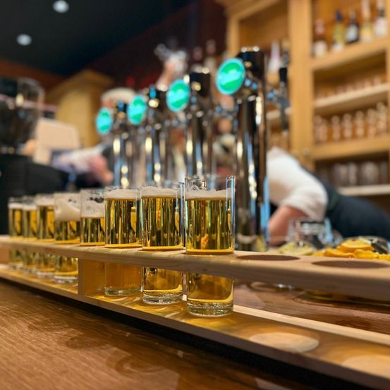 An der Bartheke sind mehrere Gläser Bier auf einem Holztablett aufgereiht, mit Zapfhähnen im Hintergrund und einer Person, die hinter der Theke arbeitet.