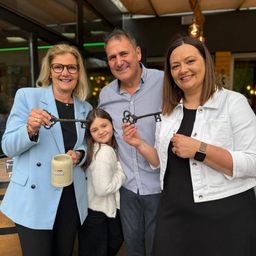 Vier Erwachsene und ein Kind lächeln in die Kamera und halten einen Schlüssel. Das Kind hält einen Becher. Sie scheinen bei einer Bandzerschneidungszeremonie zu sein.