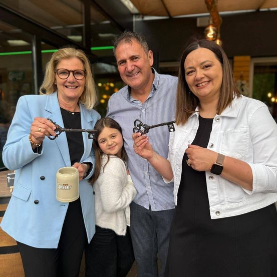 Vier Erwachsene und ein Kind lächeln in die Kamera und halten einen Schlüssel. Das Kind hält einen Becher. Sie scheinen bei einer Bandzerschneidungszeremonie zu sein.