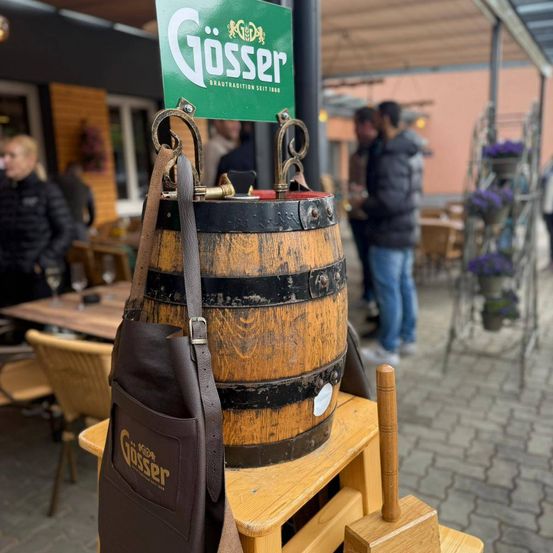 Ein hölzerner Fass mit einem Lederriemen hängt über einem Holzhocker. Eine braune Lederschürze ist in der Nähe. Das Fass hat ein grünes Schild mit der Aufschrift Gosser. Im Hintergrund stehen Menschen.