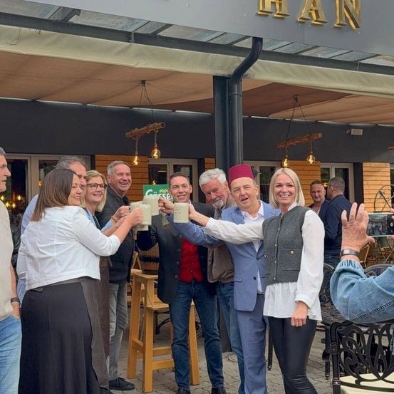 Eine Gruppe von Menschen feiert vor einem Restaurant und hält Biergläser. Eine Frau in der Mitte hebt ihre Arme. Der Name 'HAN' steht auf dem Dach.