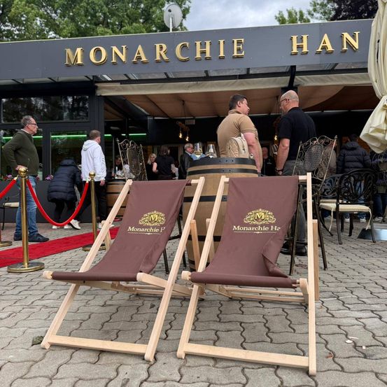 Zwei braune Monarchie Han Klappstühle stehen vor einem Restaurant. Leute stehen und sitzen herum, und ein Fass ist in der Nähe.