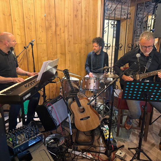 Drei Männer spielen Musik in einem Raum. Einer spielt Klavier, ein anderer Schlagzeug und der dritte Gitarre. Sie tragen alle schwarze Hemden und Brillen.