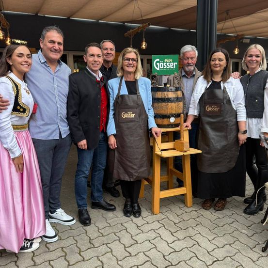 Eine Gruppe von Menschen steht um einen Fass herum, zwei Frauen in Schürzen halten dessen Griffe, ein Mann in einem Anzug lächelt und eine Frau in einem traditionellen Kleid posiert.