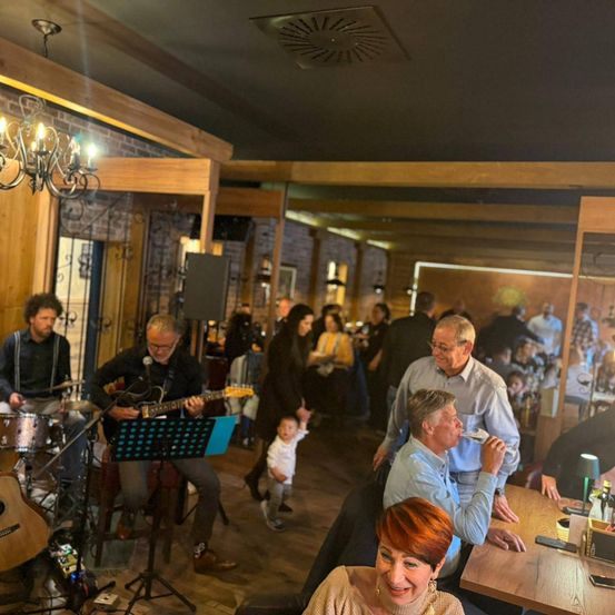 Eine Band spielt in einem lebhaften Pub mit Holzinterieurs und Ziegelwänden. Ein Publikum sitzt und steht herum, einige genießen Getränke und andere beobachten die Musiker.