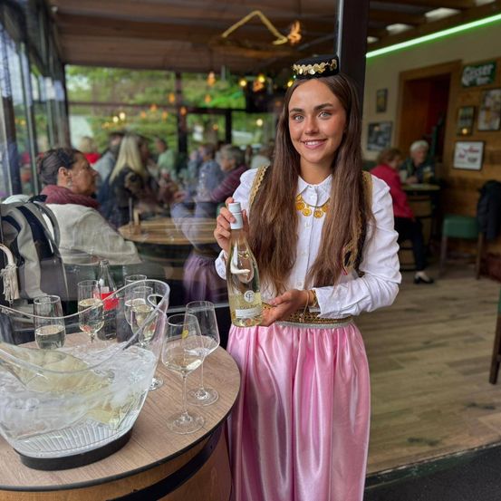 Eine Frau in einem rosa Kleid und traditioneller Kopfbedeckung hält eine Flasche Wein in einem Restaurant. Ein Weinkrug und Gläser stehen auf dem Tisch. Mehrere Personen sitzen im Hintergrund.