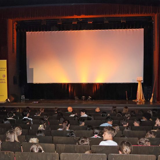 Ein großes Theater mit einer weißen Leinwand und einem Publikum, das in Reihen sitzt. Die Bühne ist leer und der Raum ist schwach beleuchtet. Ein gelber Banner ist auf der linken Seite der Bühne sichtbar.