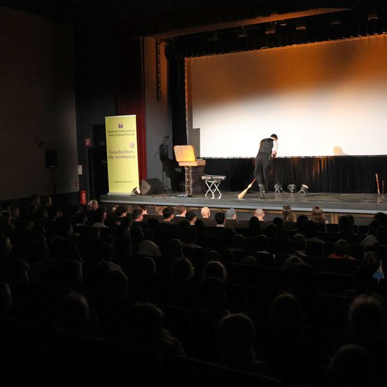 Ein Mann fegt mit einem Besen die Bühne eines Theaters, während ein Publikum zuschaut.