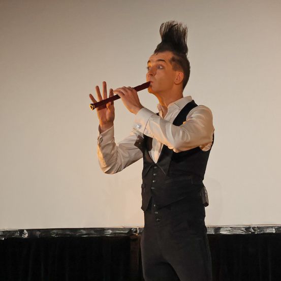 Ein Mann mit spitzen Haaren spielt auf der Bühne eine Holzflöte und trägt ein weißes Hemd und eine schwarze Weste.