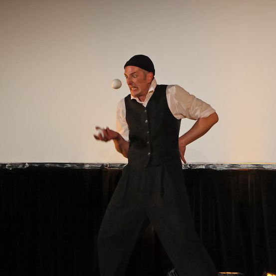 Ein Mann in einem schwarzen Vest und Hut jongliert mit einer weißen Kugel in der Luft auf der Bühne.