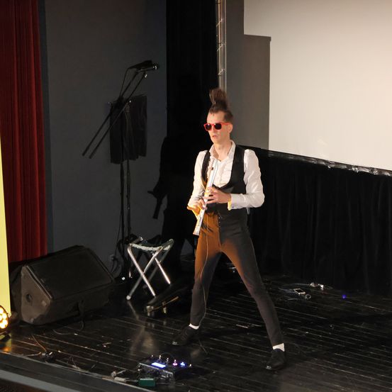 Eine Person auf der Bühne mit roten Gläsern und einem weißen Hemd spielt Gitarre. Die Bühne ist schwarz mit einem roten Vorhang und einem Mikrofon.