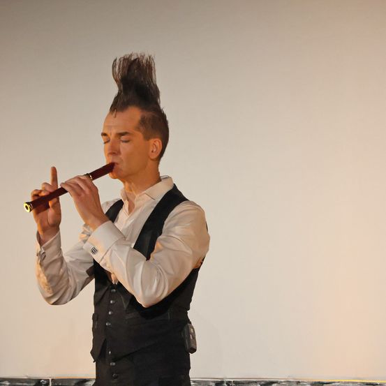 Ein Mann mit gespickten Haaren spielt eine Flöte auf der Bühne. Er trägt ein weißes Hemd und eine schwarze Weste. Der Hintergrund ist schlicht und weiß.
