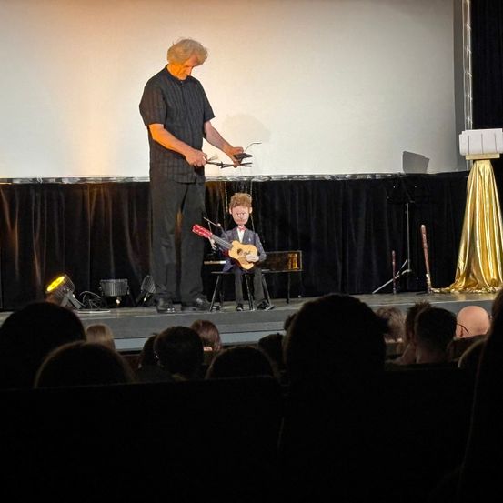 Ein Mann spielt auf der Bühne eine Gitarre, während eine kleine Person in der Nähe sitzt. Die kleine Person trägt einen Fliege und hält eine Gitarre. Scheinwerfer sind auf der Bühne.