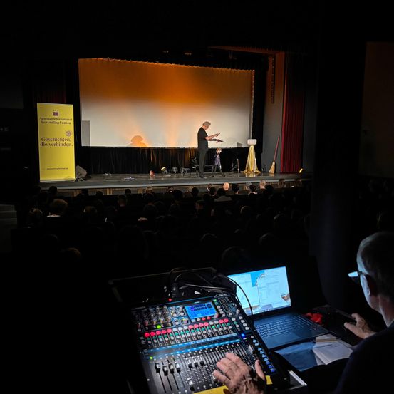 Ein Mann mit Mikrofon auf der Bühne vor einer großen Leinwand, mit einem Publikum in einem Theater. Ein anderer Mann mit Brille sitzt an einer Mischkonsole.
