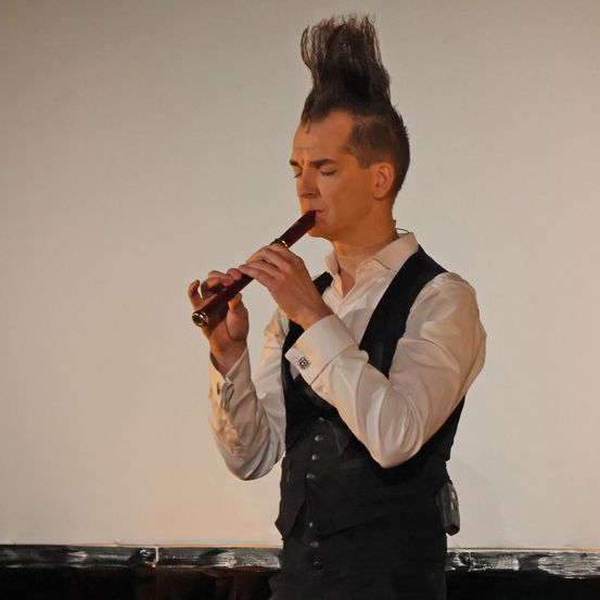 Ein Mann mit spitzem Haar spielt eine Flöte auf der Bühne. Er trägt ein weißes Hemd und eine schwarze Weste. Er schaut nach unten mit geschlossenen Augen.