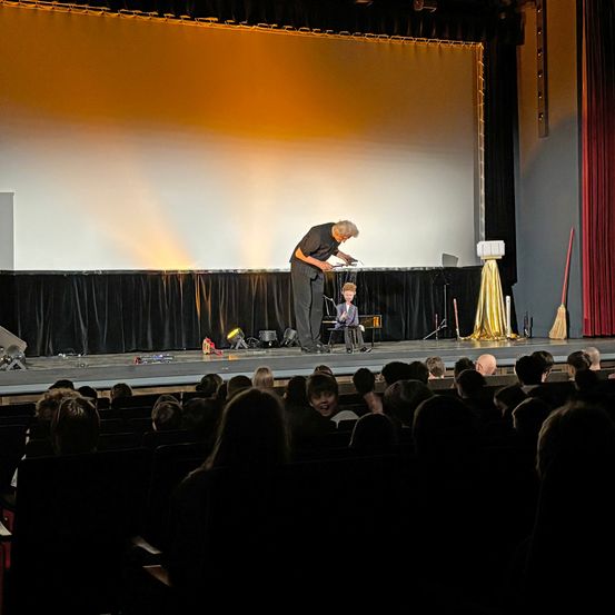 Ein Mann führt auf der Bühne einen Zaubertrick mit einem jungen Jungen vor. Das Publikum sieht im dunklen Theater zu.