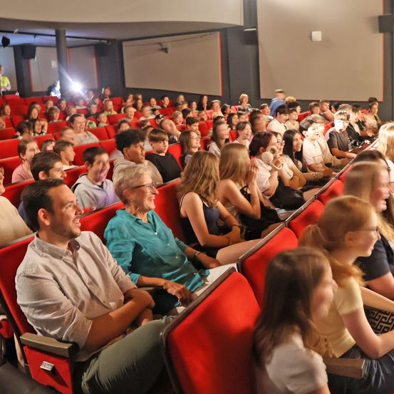 Ein überfülltes Kino mit einem vielfältigen Publikum, das sich aus Menschen unterschiedlichen Alters zusammensetzt. Einige lächeln, andere schauen aufmerksam auf die Leinwand.