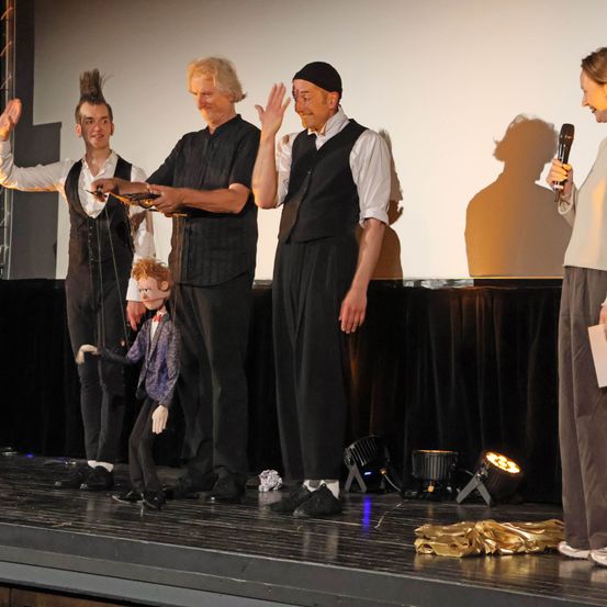 Eine Gruppe von Menschen steht mit einer Puppe auf der Bühne. Ein Mann hält einen Pokal und ein anderer ein Mikrofon. Die Puppe wird von einem der Männer gehalten.