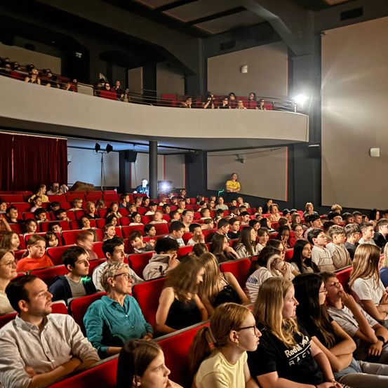 Ein Publikum aus jungen Menschen und Erwachsenen sitzt in einem Theater. Das Theater hat rote Sitze und rote Vorhänge, und einige Menschen stehen auf der Galerie.