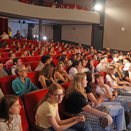 Ein volles Kino mit roten Stühlen, Leute sitzen und schauen einen Film, ein paar Leute stehen auf der Galerie und eine Person steht hinten.