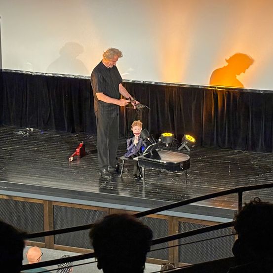 Ein Mann steht auf einer Bühne mit einer kleinen Puppe neben einem Klavier, unter Scheinwerfern, mit einem Schatten auf der Kulisse. Ein Publikum sitzt davor und schaut zu.