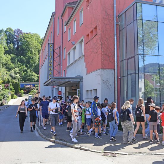 Eine Reihe von Menschen, wahrscheinlich Schüler, geht auf dem Bürgersteig vor einem Gebäude namens Kulturhaus, mit Bäumen und Bergen im Hintergrund.