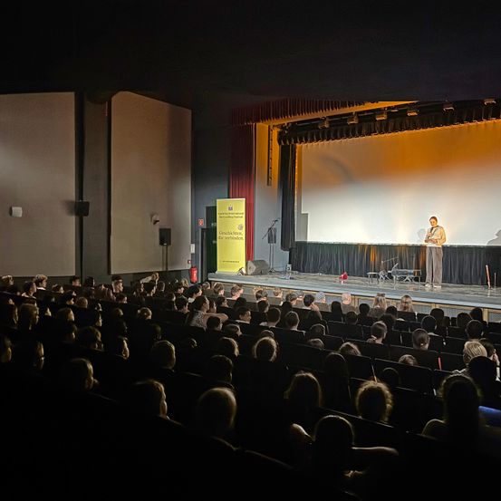 Ein Mann steht auf der Bühne eines Theaters, vor einem Publikum. Hinter ihm befindet sich ein gelbes Schild und ein roter Vorhang mit einer Leinwand auf der Bühne.