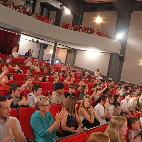 Ein mit verschiedenen Personen gefülltes Auditorium klatscht, einige in formeller Kleidung, in einem Theater mit roten Sitzen und hellen Lichtern.