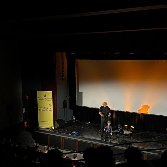 Ein Mann steht mit einer Marionette auf der Bühne und führt ein Puppenspiel vor einem Publikum vor, mit einer großen Leinwand dahinter und einem Banner auf der linken Seite.