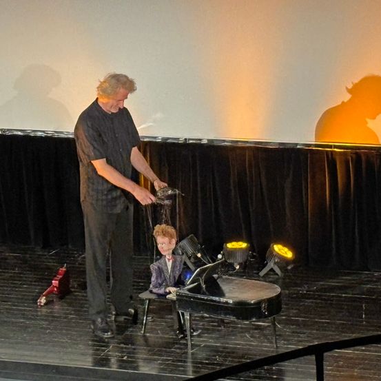 Ein Mann auf der Bühne gießt Wasser über eine kleine Puppe, die an einem Klavier sitzt, während Scheinwerfer die Szene beleuchten.