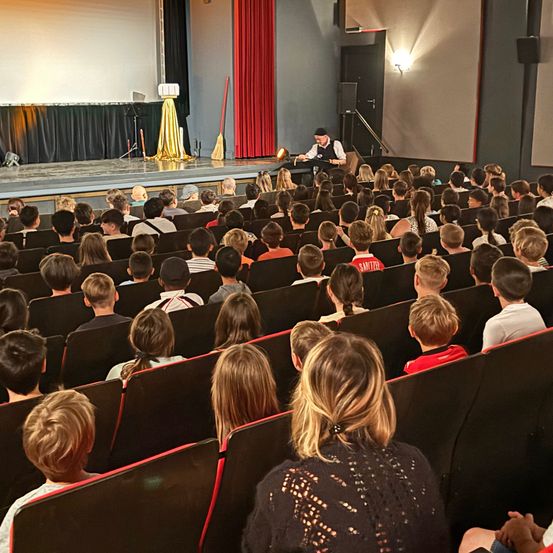Eine Theateraufführung läuft. Ein Mann sitzt auf der Bühne vor einem Publikum. Die Bühne hat Requisiten und eine Leinwand. Das Publikum sitzt in Reihen und schaut aufmerksam zu.