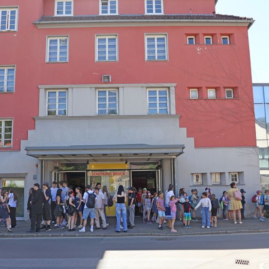 Viele Menschen, einschließlich Kinder, warten vor einem roten Gebäude, möglicherweise einem Museum. Sie sind in der Nähe des Eingangs versammelt, mit Schildern, die Stadtkino Bruck und Kleine lesen.
