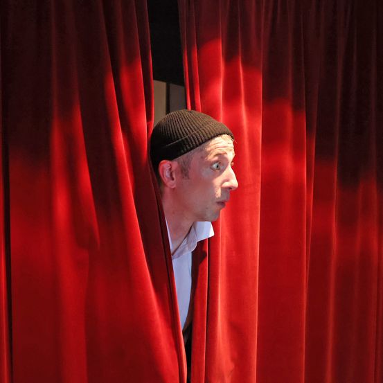 Ein Mann in einer schwarzen Mütze schaut überrascht hinter roten Theatervorhängen hervor.
