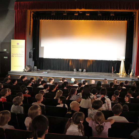 Ein überfülltes Theater mit Bühne, einer großen Leinwand und vielen Kindern beim Zuschauen. Ein Schild mit 'Geschichten die verbinden' steht auf der linken Seite der Bühne.