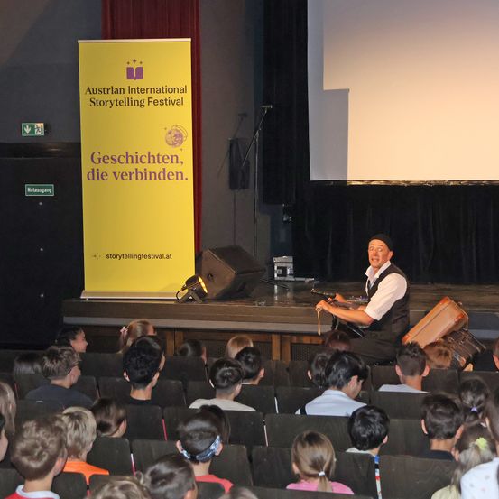 Ein Geschichtenerzähler tritt in einem Theater mit einem Publikum von Kindern auf. Die Bühne hat eine Kulisse und ein Banner mit einem Logo.