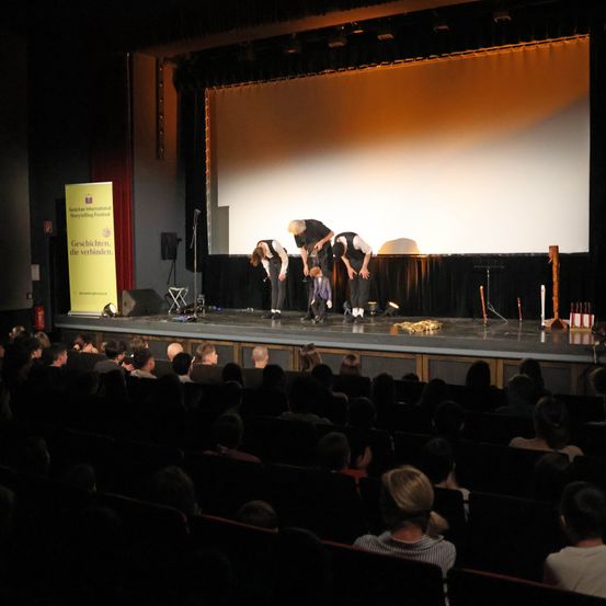 Eine Theateraufführung findet statt, bei der drei Personen auf der Bühne stehen, eine davon kniet. Die Bühne hat ein Mikrofon, einen Lautsprecher und ein gelbes Banner mit Text. Das Publikum sitzt vor der Bühne.